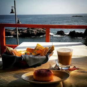 Une table avec une assiette de nourriture, un verre de bière et un panier de collations, le tout placé près d’une rambarde donnant sur la mer.
