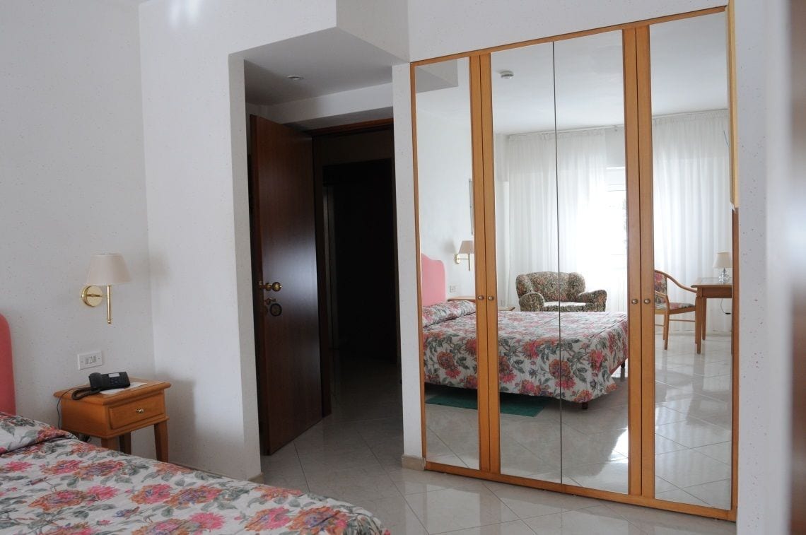 Una camera d'albergo con un'armadio in legno, un letto con lenzuola a motivi floreali, un comodino con una lampada e una sedia nell'angolo.