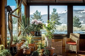 Una habitación acogedora con un amplio ventanal con vistas a un paisaje montañoso nevado, con varias plantas en macetas y muebles de mimbre.