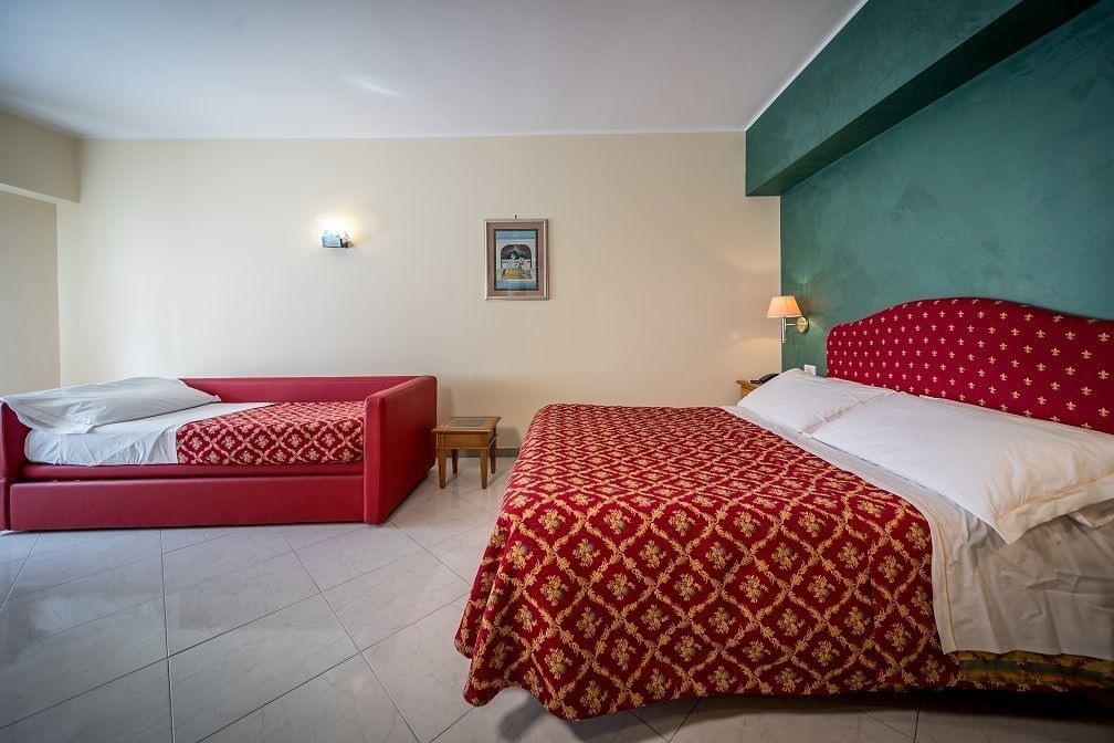 Una habitación de hotel con cama y sofá rojos, una mesita pequeña y una lámpara fijada a la pared.
