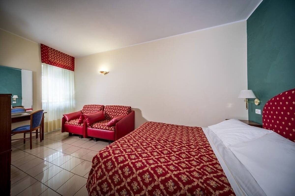 Une chambre d’hôtel avec un lit, un canapé rouge, un bureau avec une chaise et une fenêtre avec des rideaux.