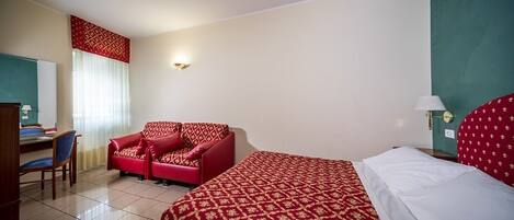 Habitación de hotel con cama, sofá rojo, escritorio y silla, y ventana con cortinas.