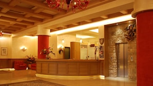 A hotel lobby with a reception desk, a red pillar, a stone wall, an elevator, and a chandelier.