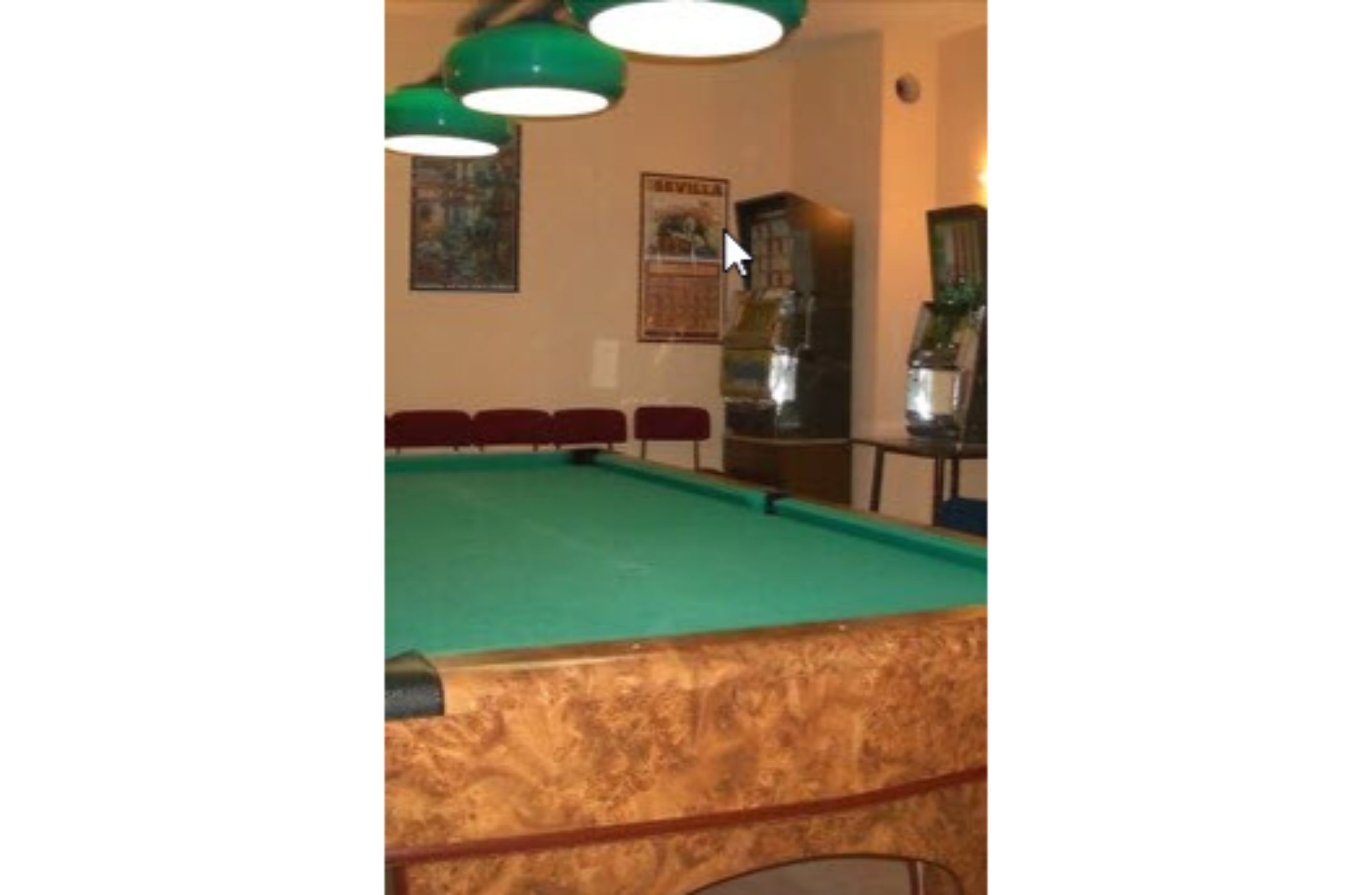 Una mesa de billar con fieltro verde, rodeada de sillas rojas, en una habitación con lámparas colgantes verdes y un cartel en la pared.