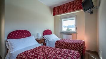 Una stanza con due letti, ognuno con copripiumino rosso fantasia e cuscini bianchi. È presente un comodino con una lampada, una finestra che dà sulle montagne e un televisore a parete.