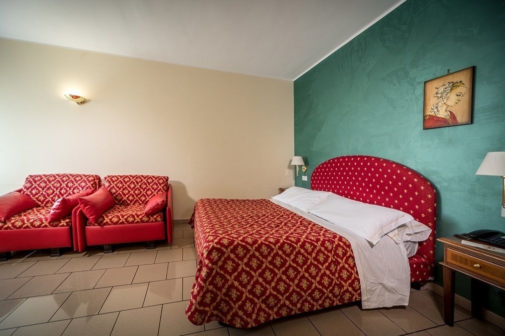 A hotel room with a bed, a red sofa, a nightstand, and a wall-mounted lamp.