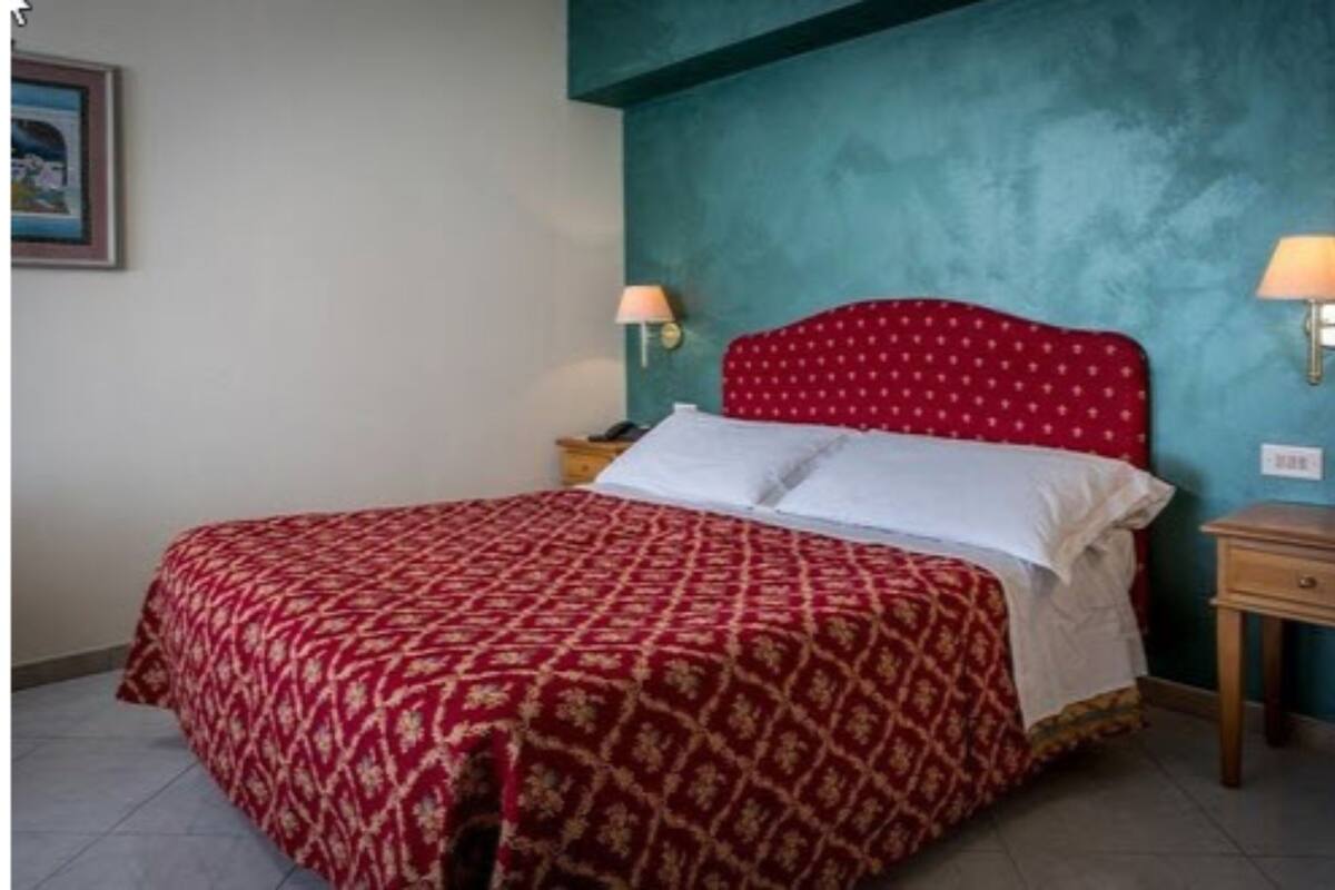 Une chambre à coucher avec une couvre-lit à motifs rouges, une tête de lit rouge, deux lampes de chevet et un mur d’accent de couleur turquoise.