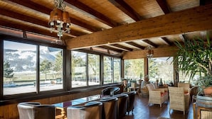 A spacious indoor area with wooden beams, large windows offering mountain views, and comfortable seating arrangements.