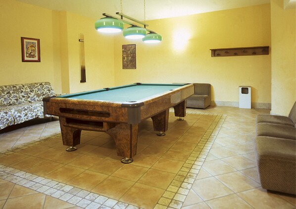 Una mesa de billar en una habitación con piso de baldosas, un sofá y un cubo de basura.
