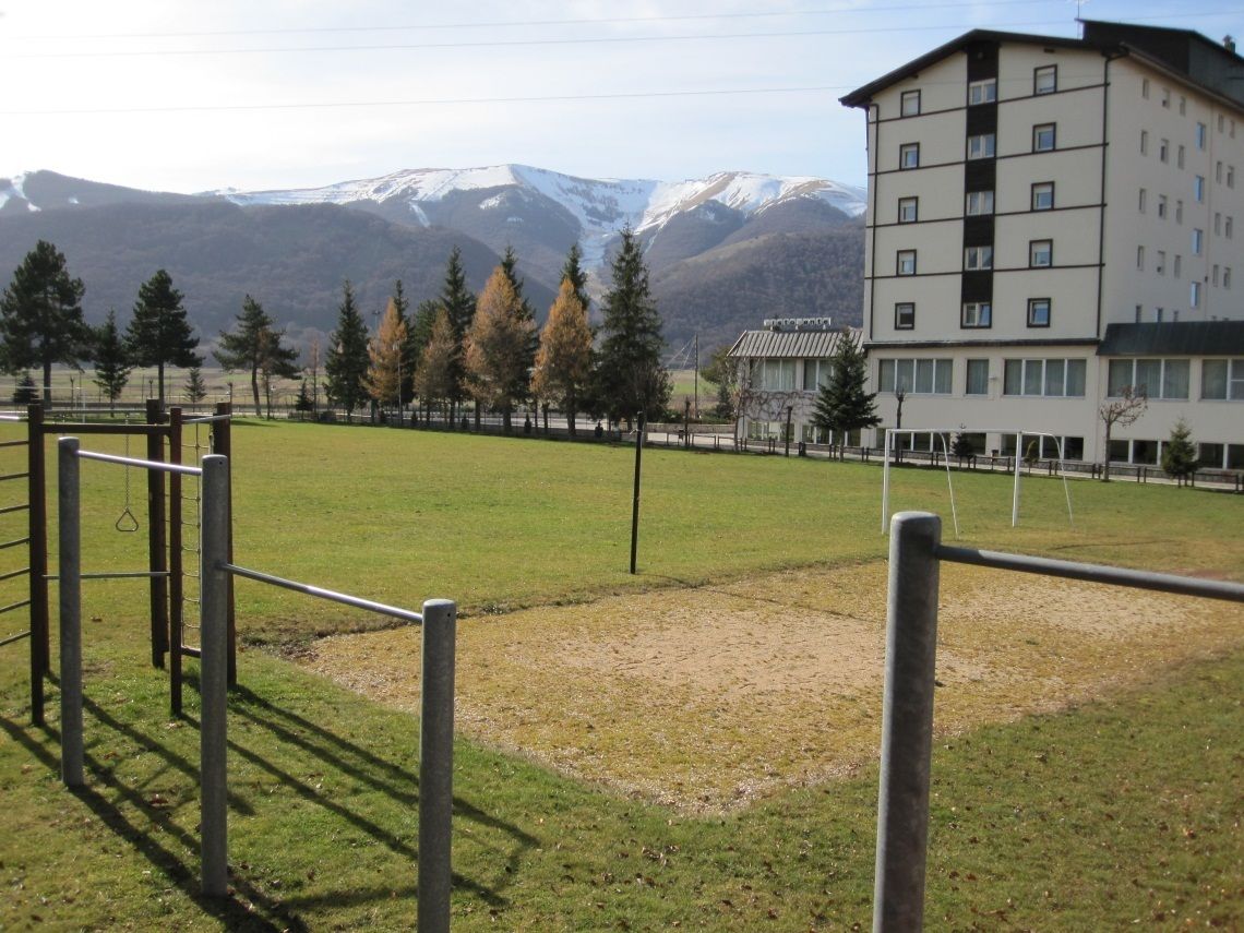 Un campo erboso con attrezzature da gioco in metallo, un edificio con diverse finestre e montagne innevate sullo sfondo.