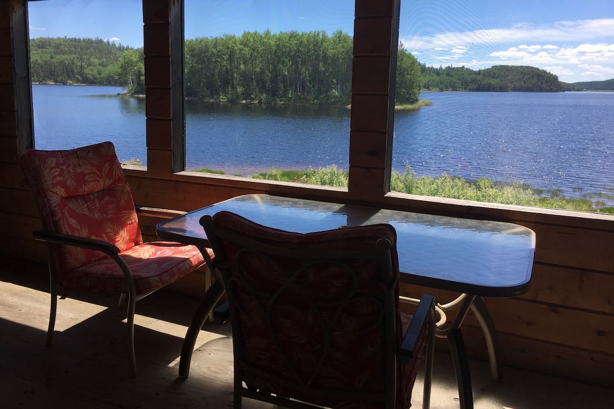 A cabin with a table and two chairs facing a large window overlooking a lake and forest.
