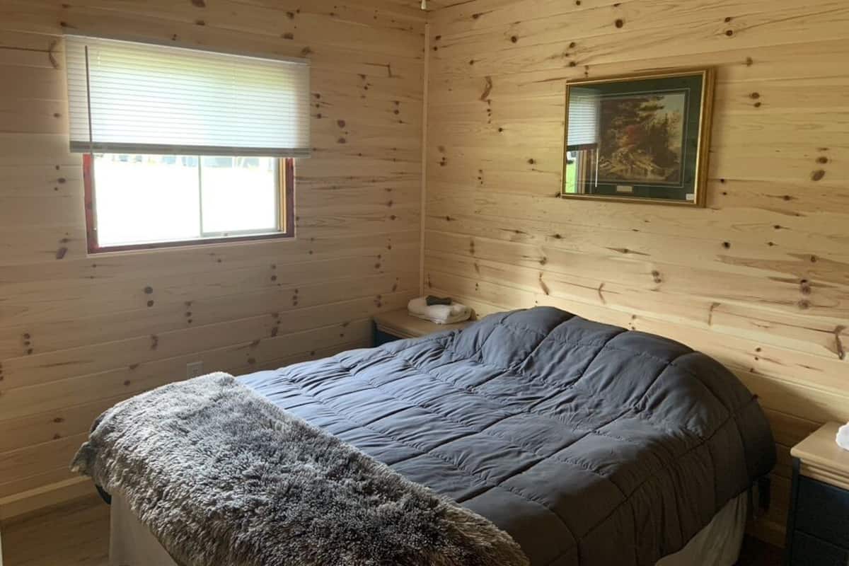 A wooden cabin bedroom with a bed, a window with blinds, a framed picture on the wall, and a small bedside table.