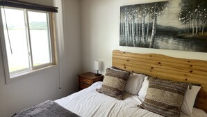 A bedroom with a wooden headboard, a large painting of a forest, a window with a view, and a bedside table with a lamp. - Air-Dale Fishing and Hunting (Unorganized North Algoma)