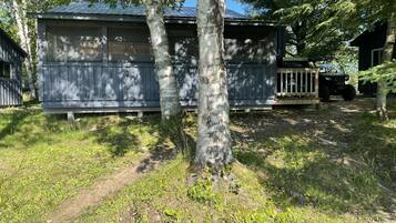 Une petite cabane à un seul étage, dotée d’une véranda grillagée, entourée d’arbres et bordée d’un sentier de terre.