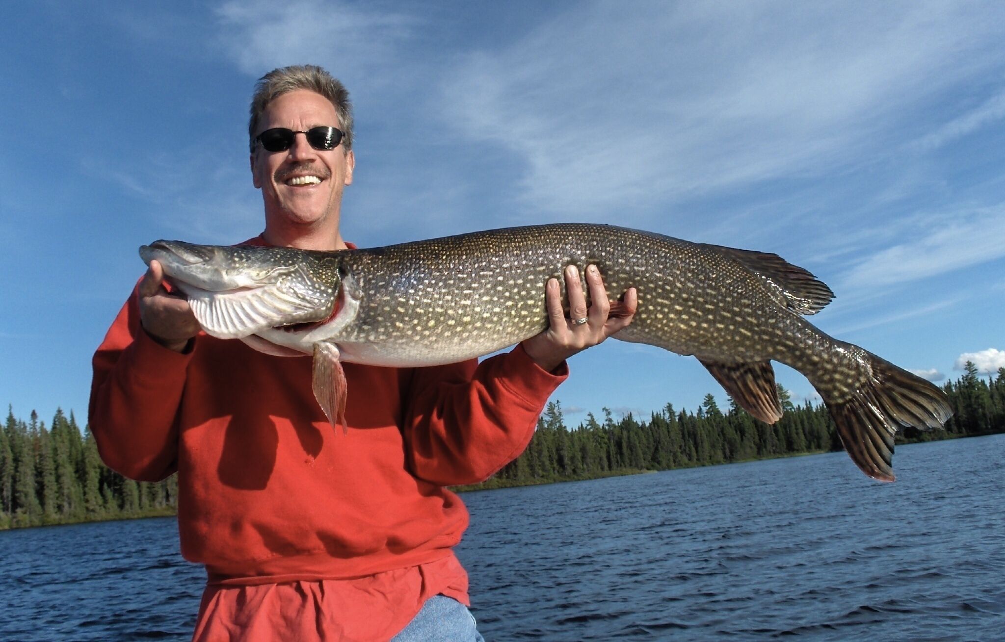 A person holding a large fish by a lake with trees in the background.