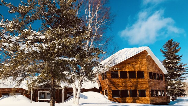 A wooden cabin with a sloped roof, surrounded by snow-covered trees and a clear blue sky. - Air-Dale Fishing and Hunting (Unorganized North Algoma)