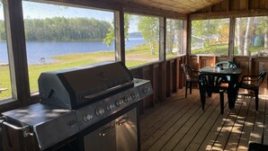 A covered outdoor area with a grill, a table with chairs, and a view of a lake. - Air-Dale Fishing and Hunting (Unorganized North Algoma)