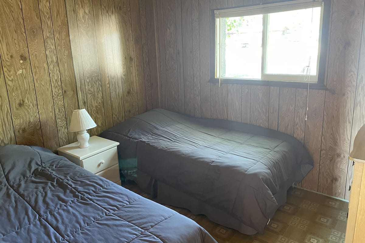 A small, single-room cabin with wooden walls, a window, a bed, and a nightstand.