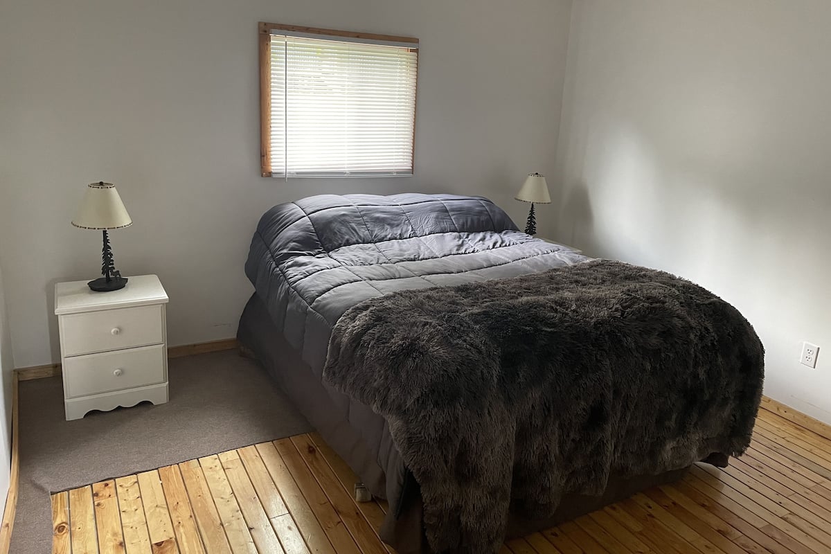 A bedroom with a bed, two bedside tables with lamps, a window, and a wooden floor.