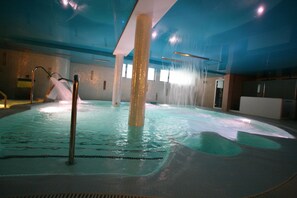Una piscina interior con múltiples chorros de agua, una columna central y suelo de baldosas.