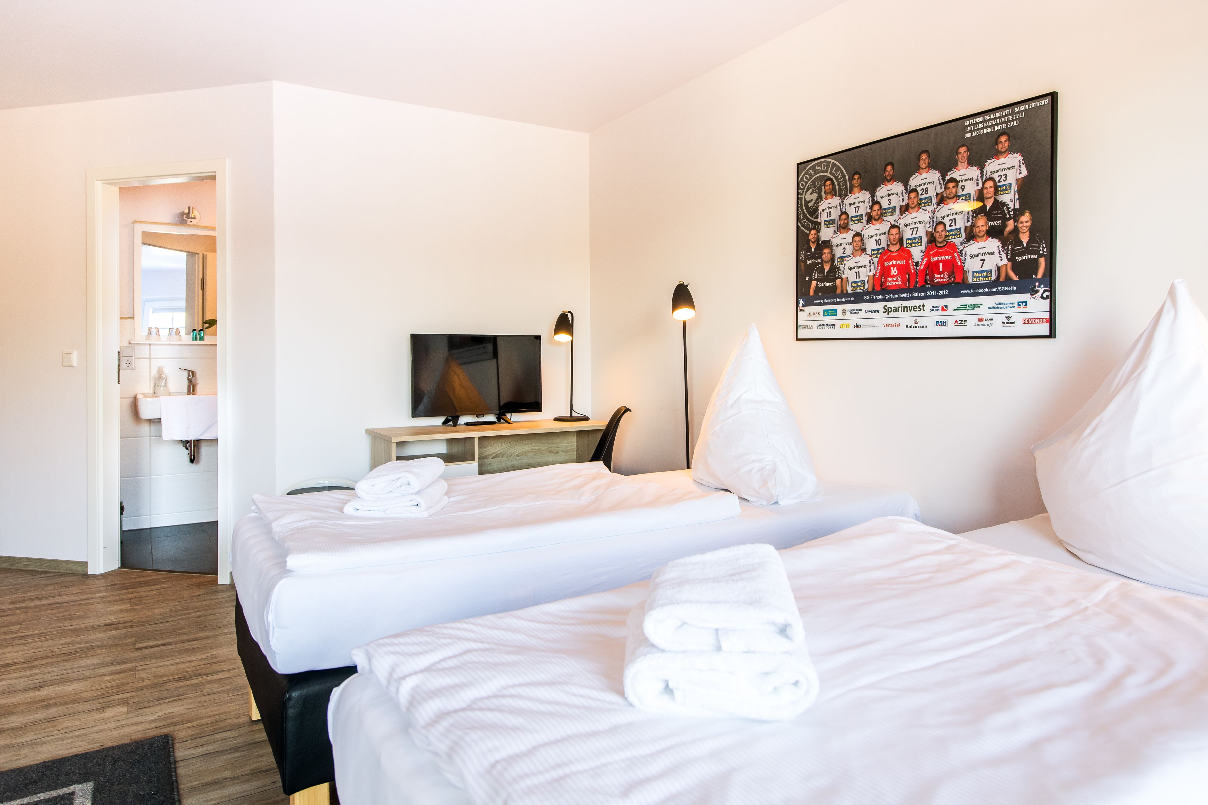A hotel room with two beds, a TV, a desk, and a framed team photo on the wall.