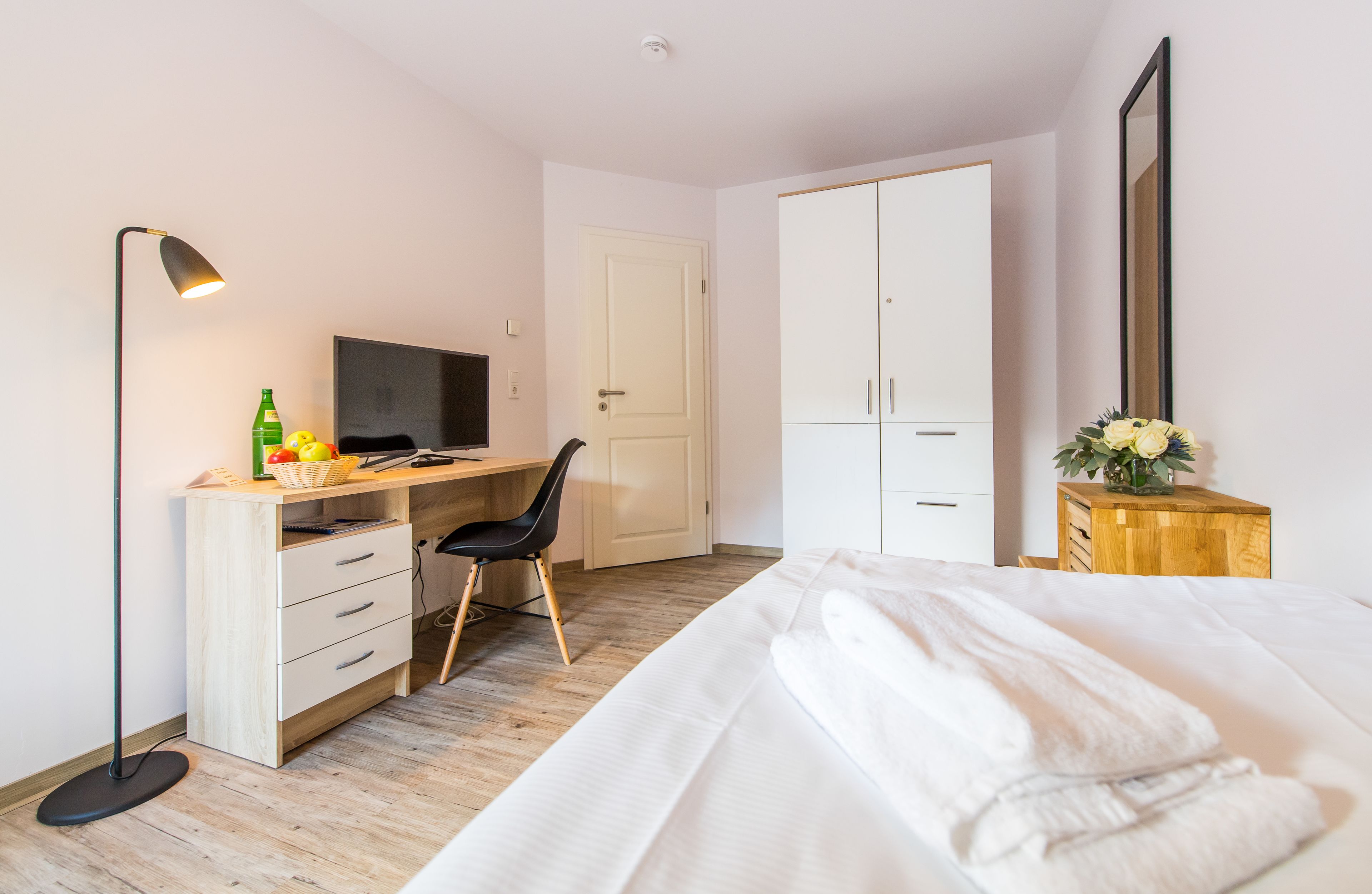 A bedroom with a bed, a desk with a chair, a TV, a wardrobe, and a bedside table with a vase.