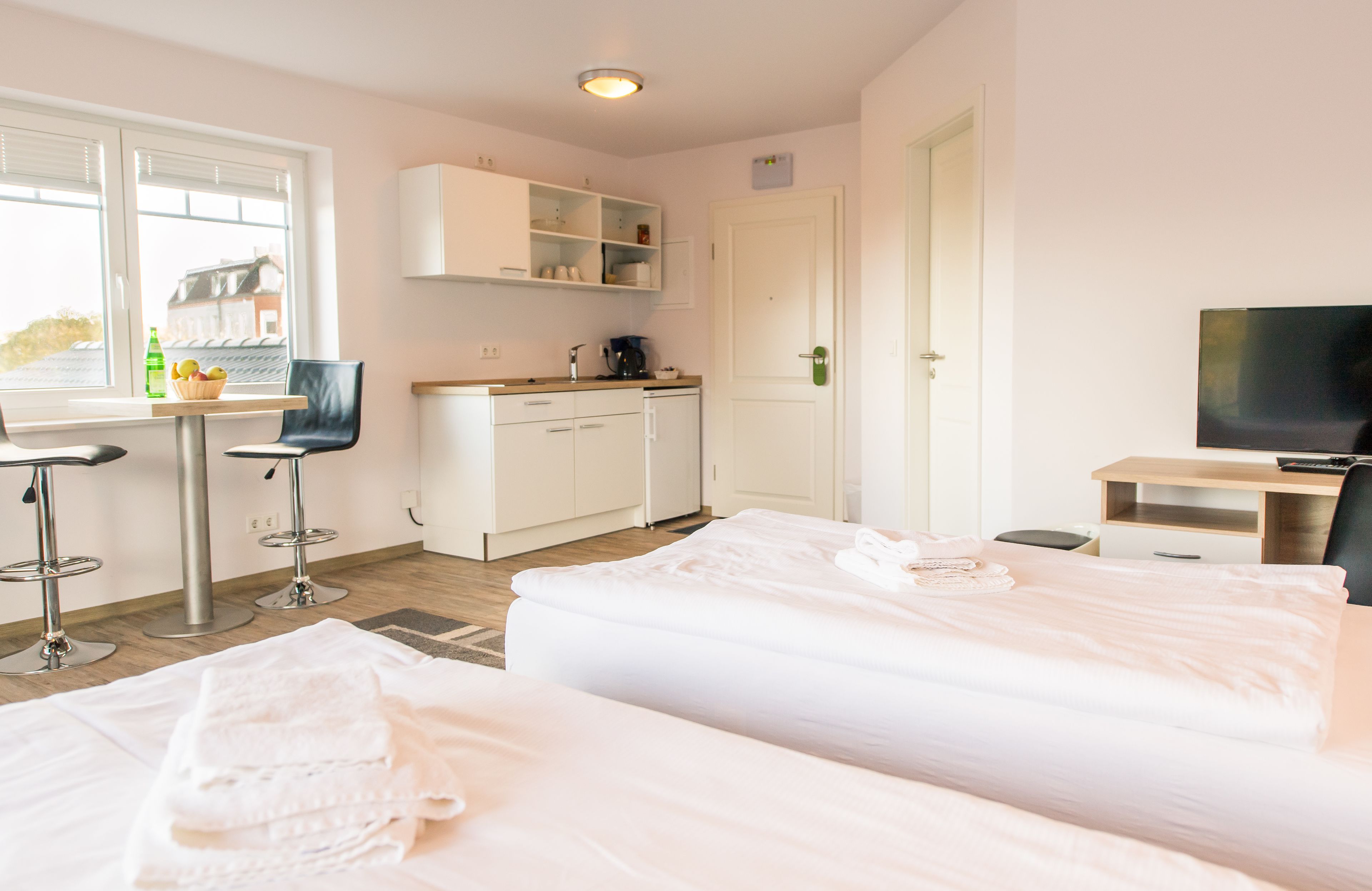 Ett hotellrum med två sängar, ett litet kök, en matplats med bord och stolar, en TV och ett fönster med persienner.
