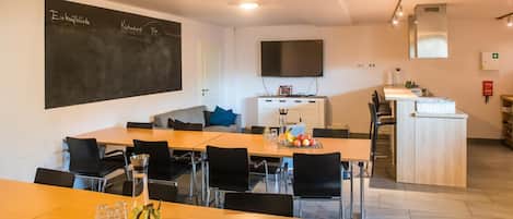 A modern dining area with a long wooden table, black chairs, and a fruit bowl. There is a chalkboard on the wall, a flat-screen TV, and a kitchen area with a sink and countertop.