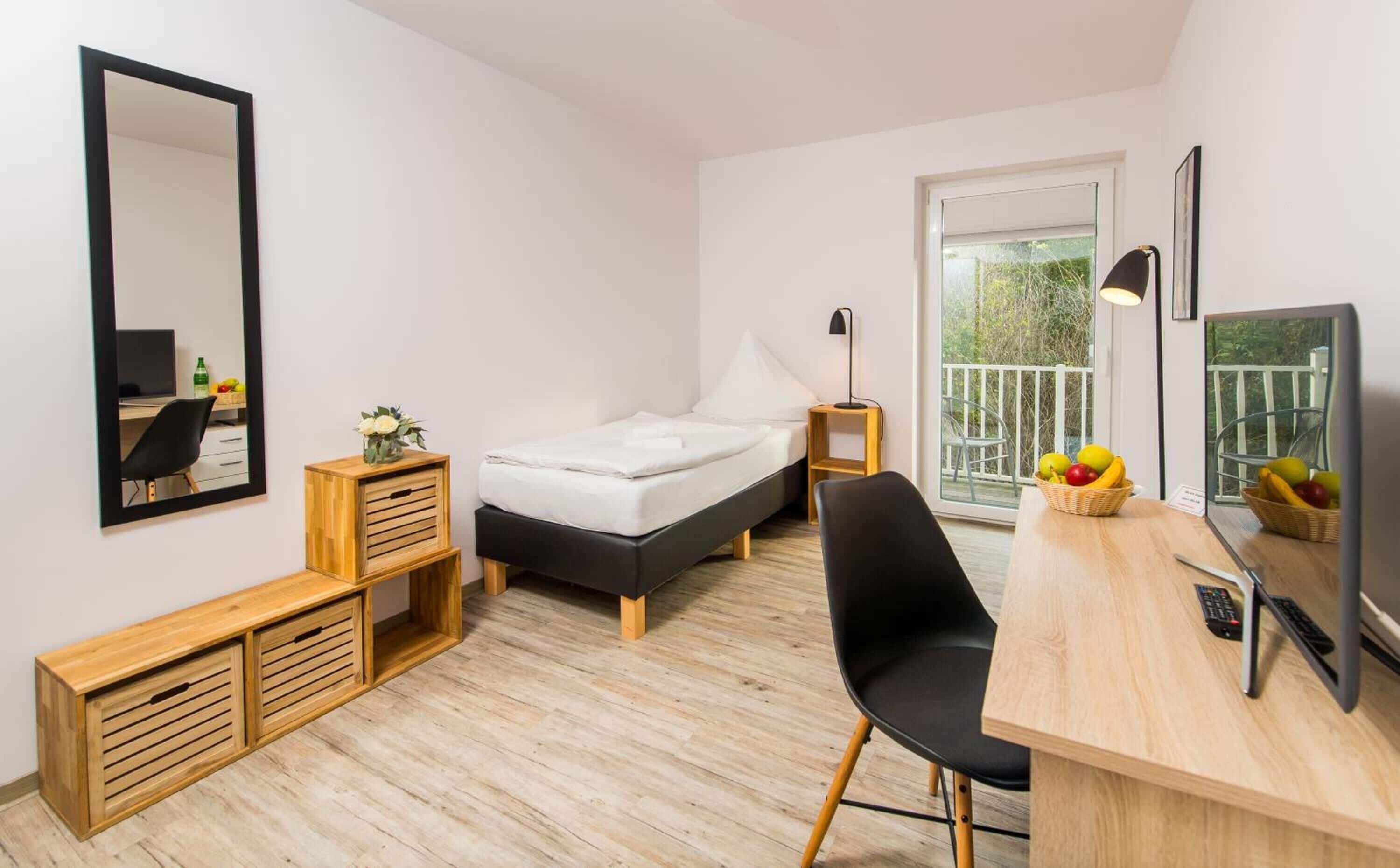 A compact bedroom with a bed, bedside table, chair, desk, and a large mirror.