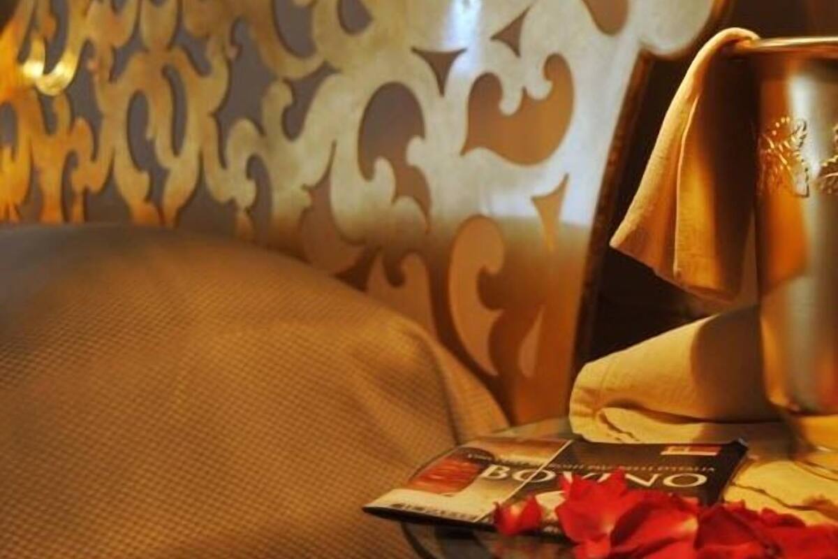 A room with a patterned headboard, a bedside table with a book and a red rose, and a lamp providing warm light.