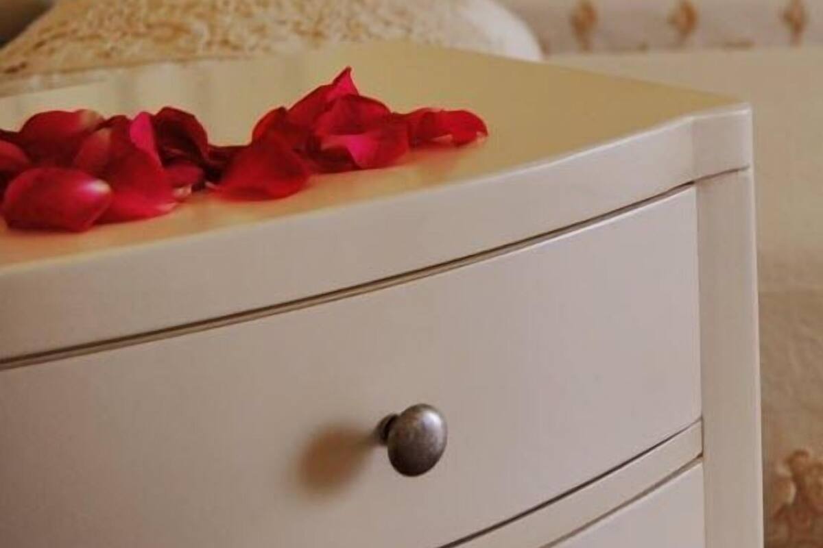 A white bedside table with a drawer and a small round knob, placed in a bedroom with a bed in the background.
