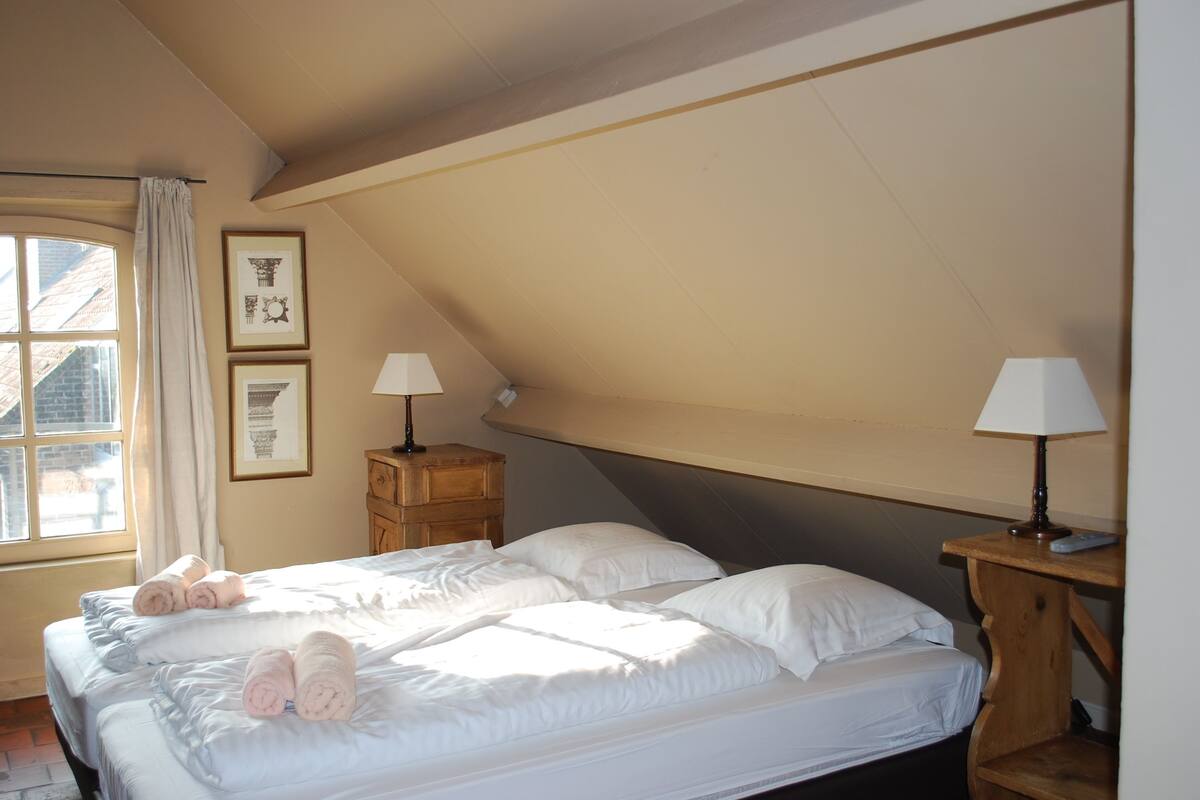 A bedroom with a large bed, two bedside tables, a window with curtains, and framed pictures on the wall.