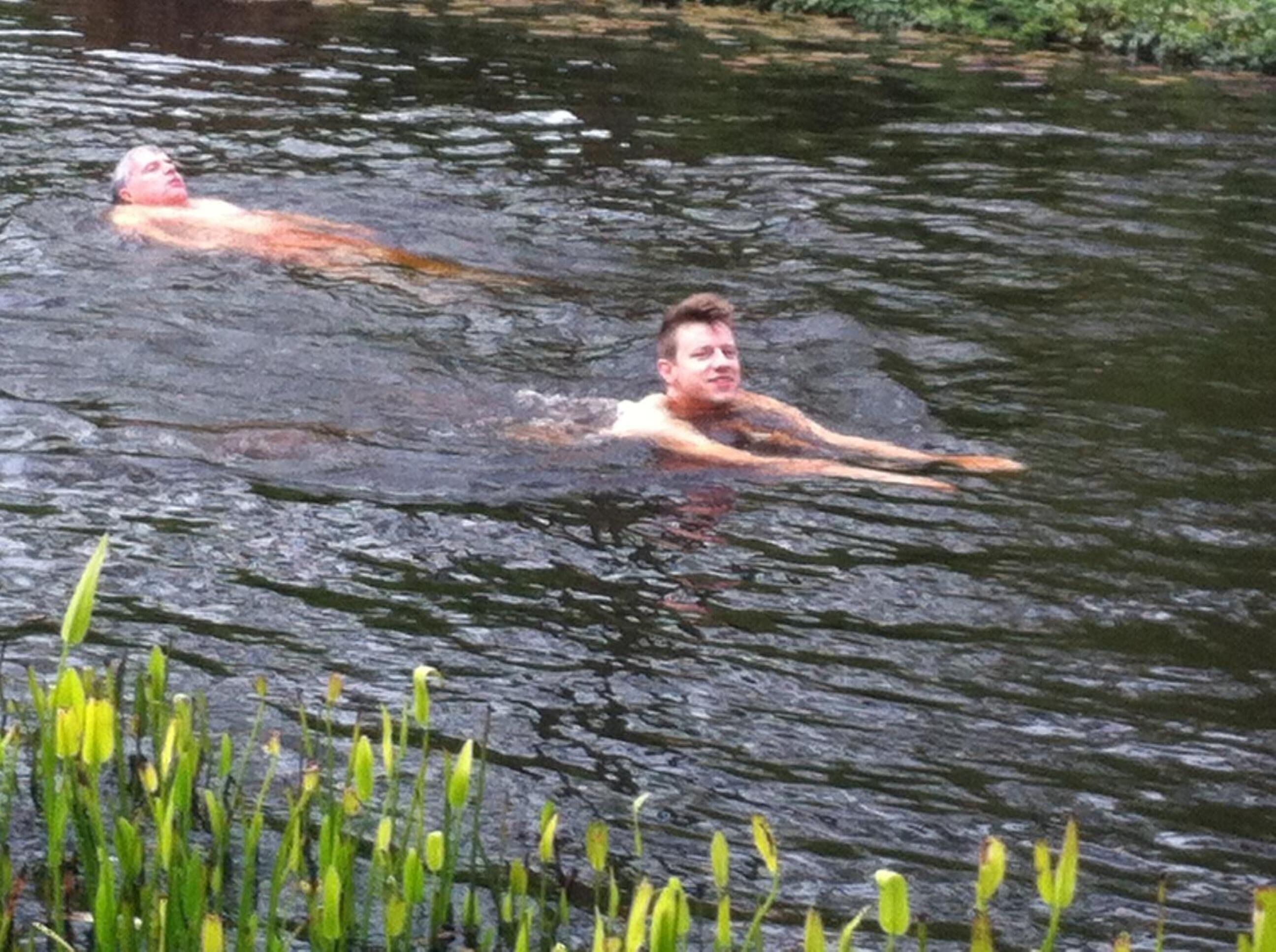 Two people swimming in a natural water body surrounded by greenery.