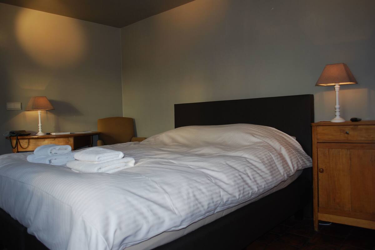 A neatly made bed with white linens and towels, a wooden nightstand with a lamp, and a wooden cabinet.
