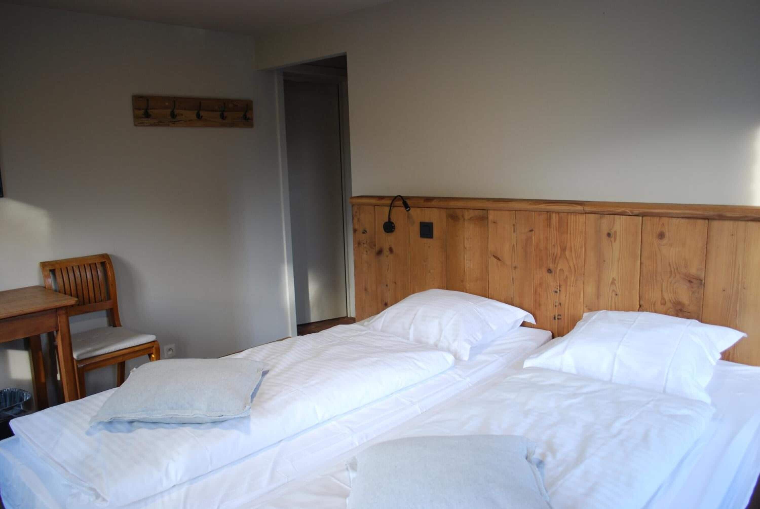 Una habitación con tablero de cama de madera, cama con ropa de cama blanca, una silla de madera y una mesita.