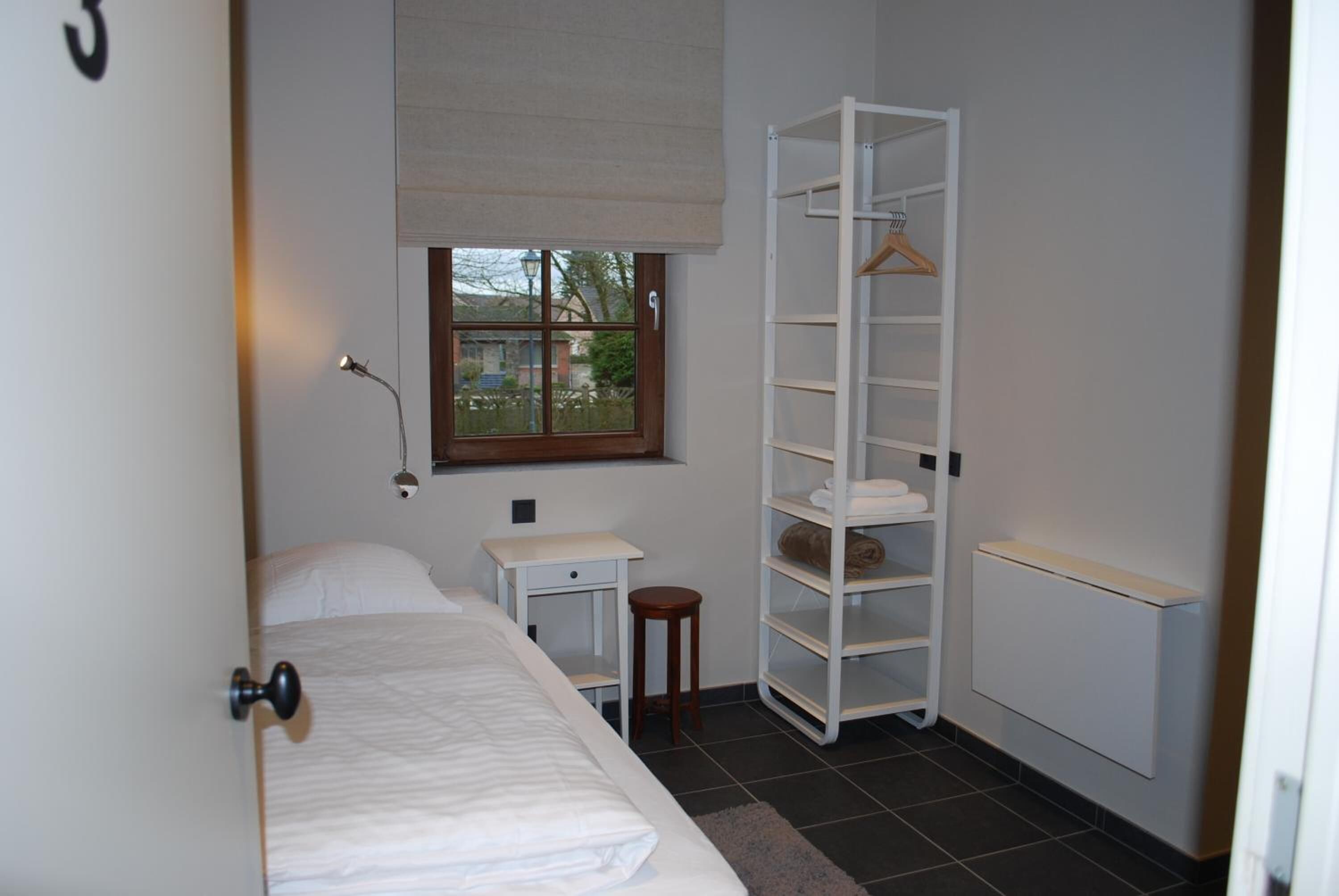 Ein Schlafzimmer mit einem Bett, einem Nachttisch, einer Lampe, einem Fenster, einer weißen Schrankwand und einer Heizung.
