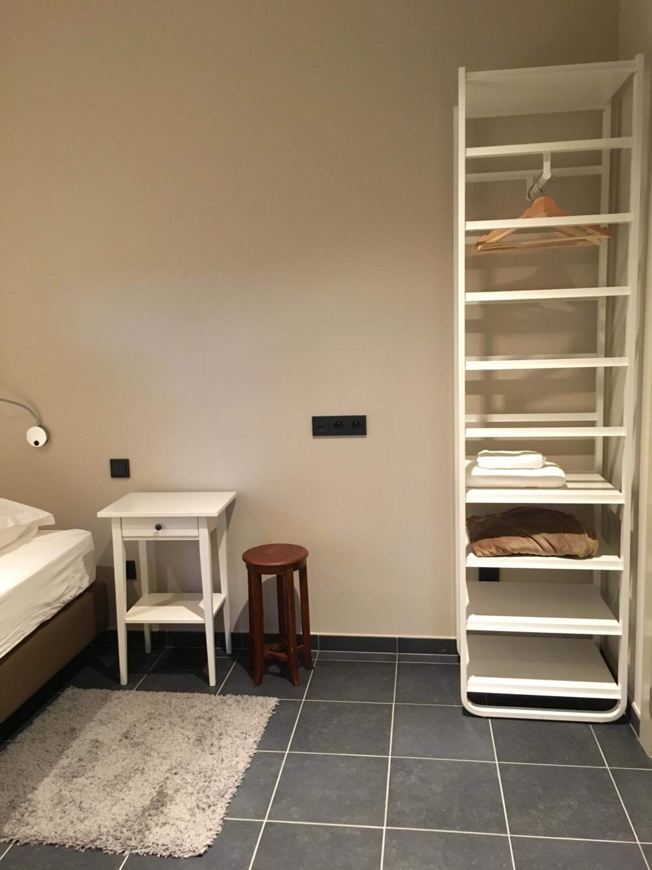 Una camera da letto con un letto, un comodino bianco, uno sgabello di legno, un ripiano portaoggetti bianco e un tappeto bianco.