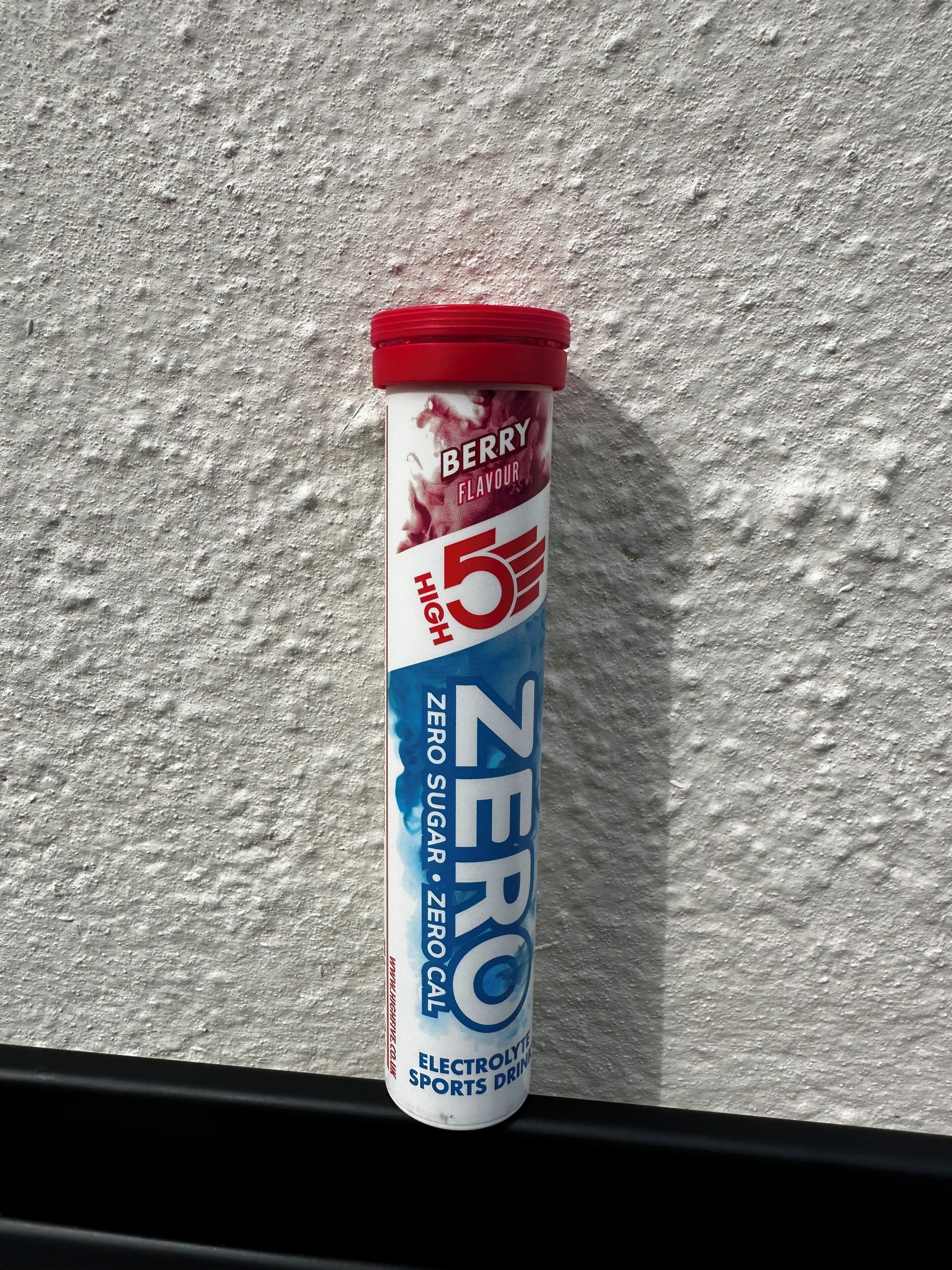A container of Zero sports drink with a red cap, labeled as Berry flavor, on a textured wall.