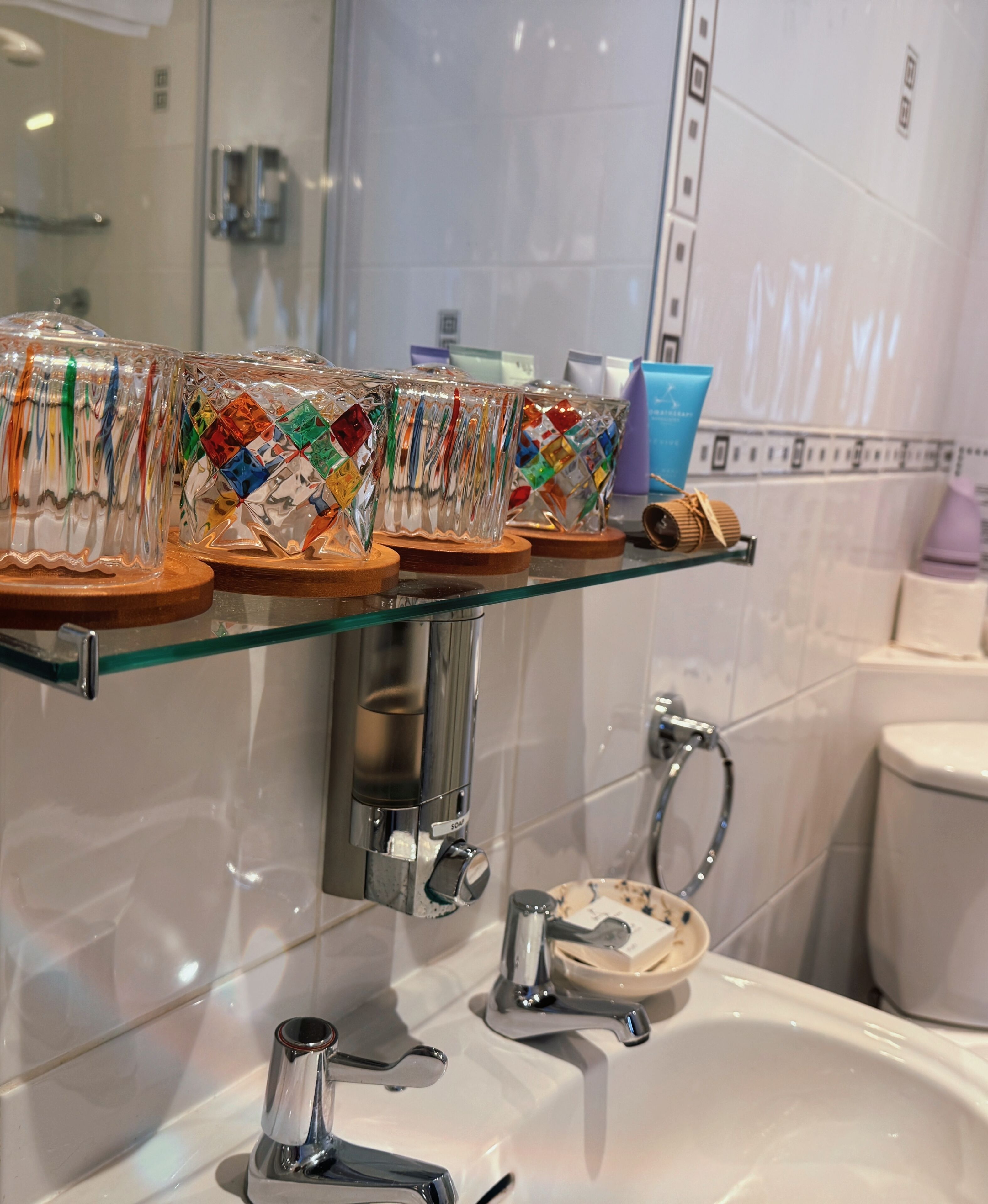 A bathroom with a glass shelf holding colorful glass containers, a white sink, and a toilet.