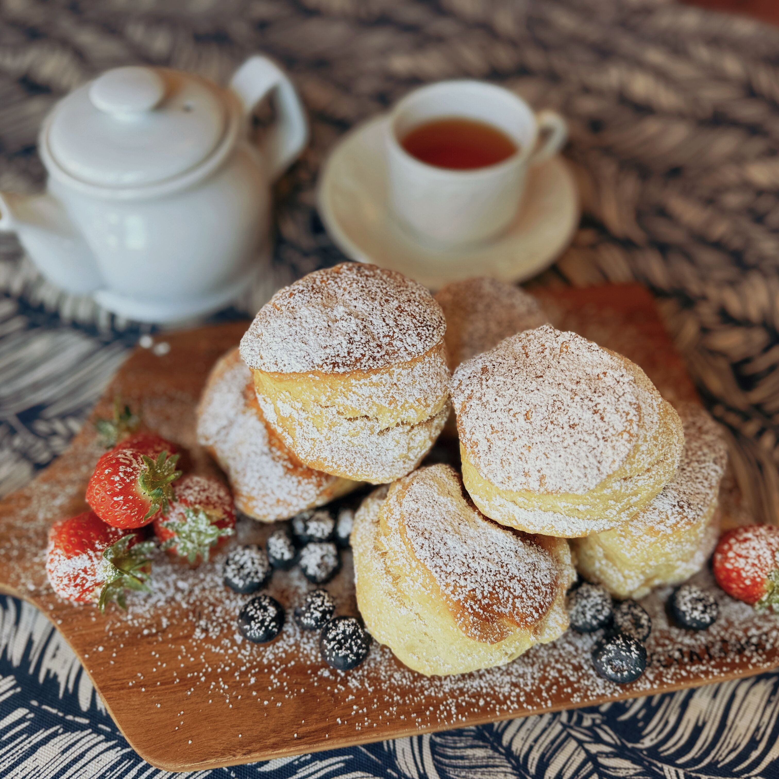 A plate of pastries dusted with powdered sugar, accompanied by a teapot and a cup of tea on a patterned tablecloth.