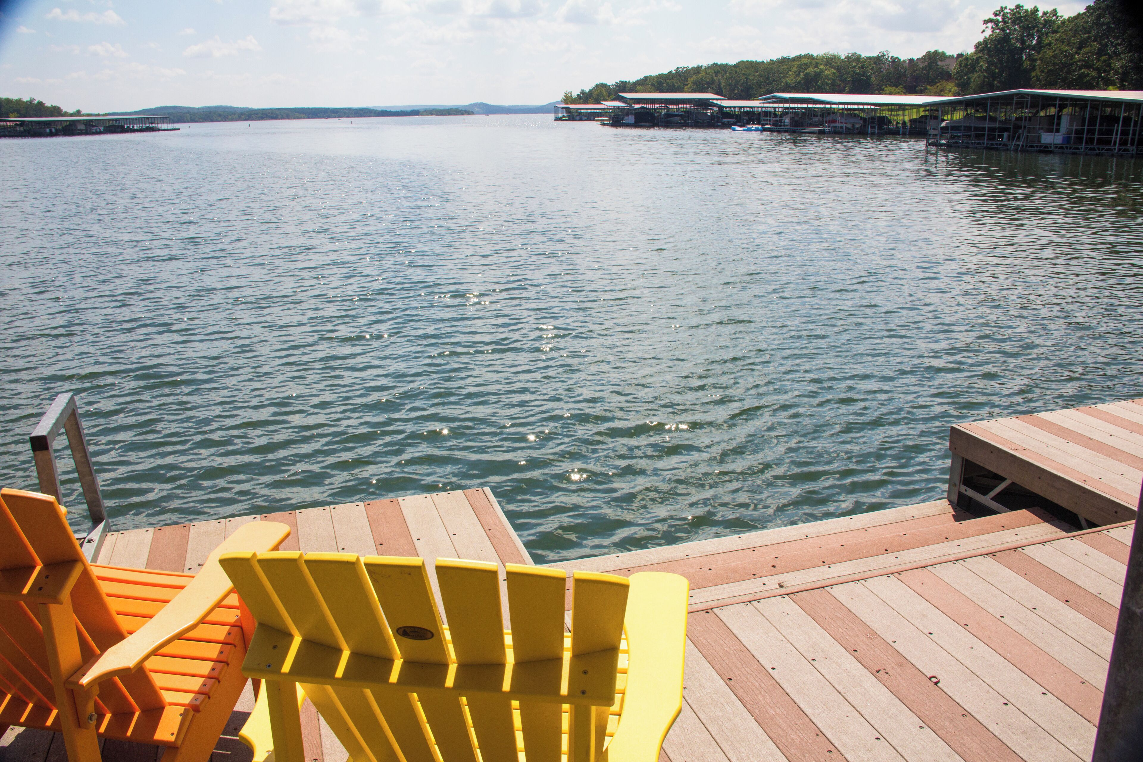 Une jetée en bois avec des chaises jaunes, donnant sur une étendue d’eau calme, une rive lointaine et un espace abrité sous un toit.