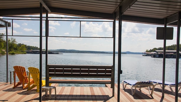 A covered dock with a bench and Adirondack chairs, overlooking a calm lake with distant land and dock structures.