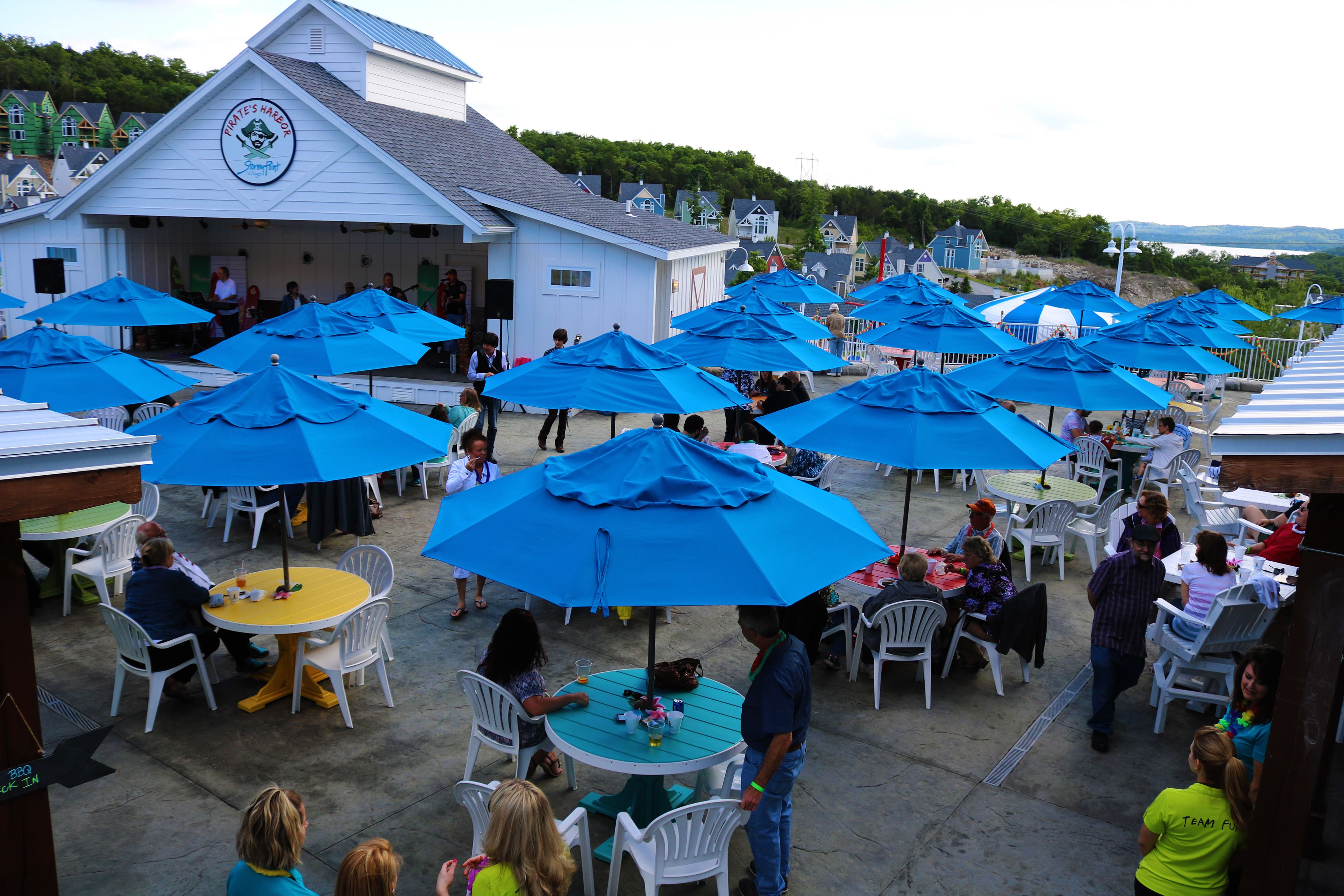 An outdoor seating area with numerous blue umbrellas and white chairs, a building with a sign, and a view of houses in the background.