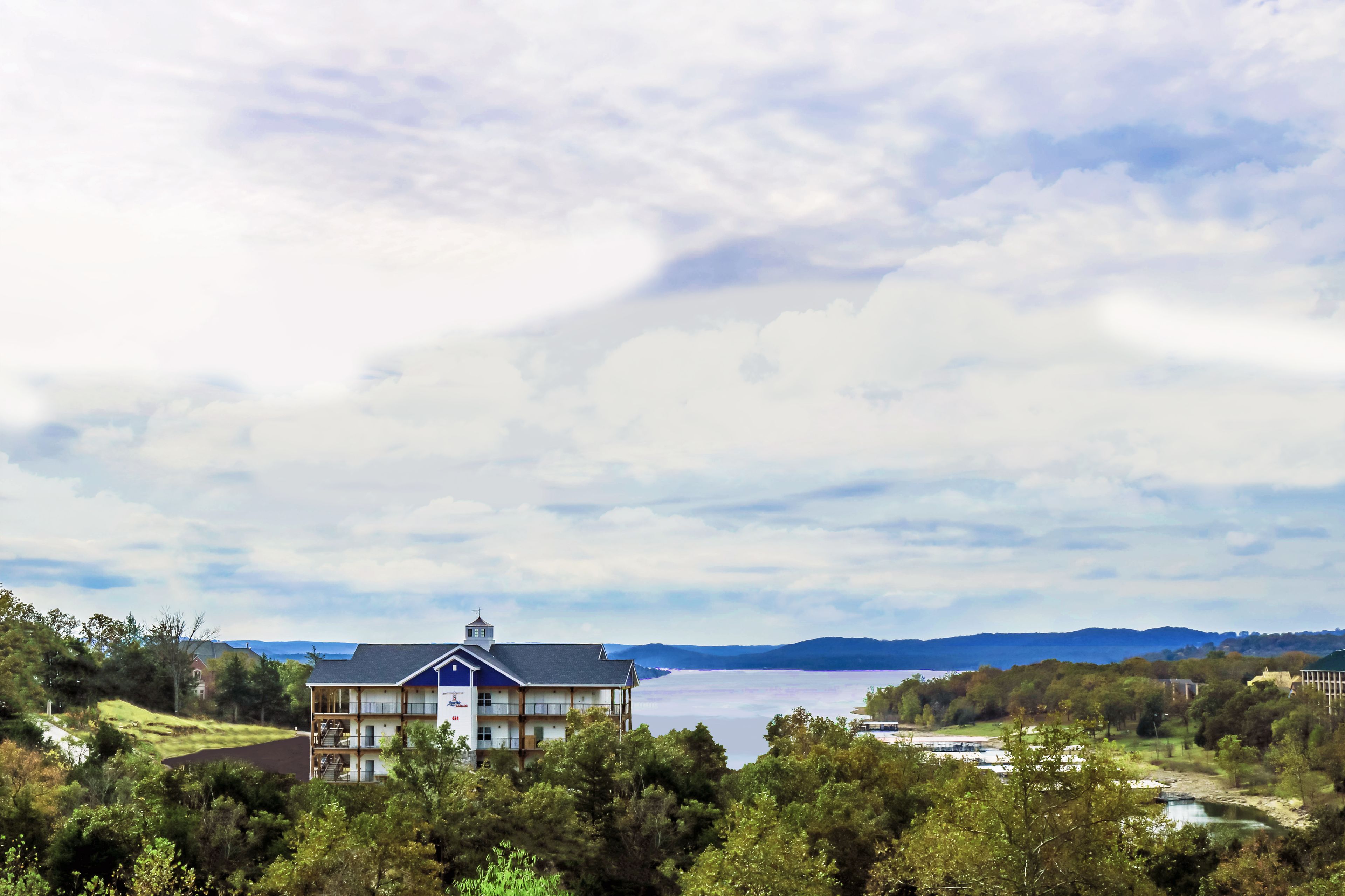 A large building with multiple floors and a prominent balcony, surrounded by lush greenery and a lake view.