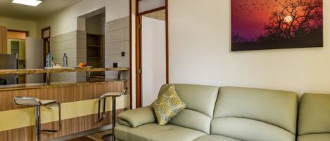 Una sala moderna con un sofá, una mesa de centro con una bandeja de frutas, una zona de cocina con una barra, y un cuadro de un atardecer.
