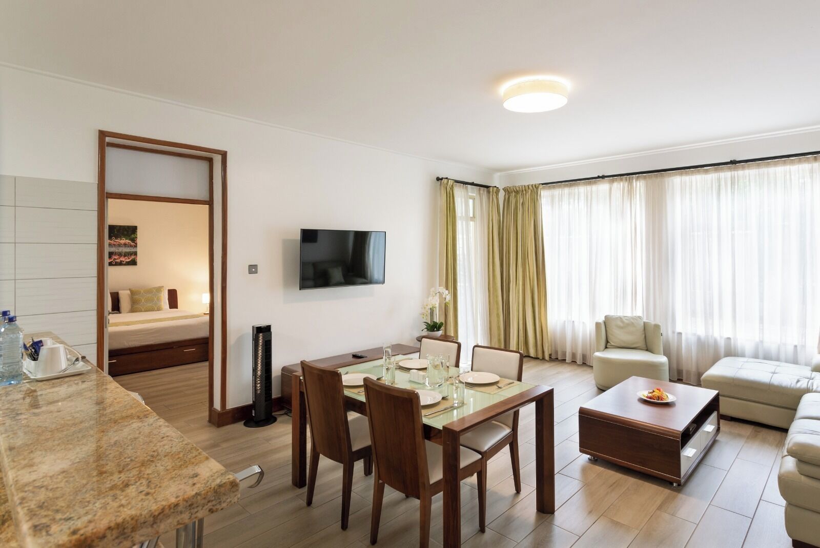 Une chambre d’hôtel comprenant un coin repas, un téléviseur fixé au mur, un lit avec une table de chevet et une grande fenêtre avec des rideaux.