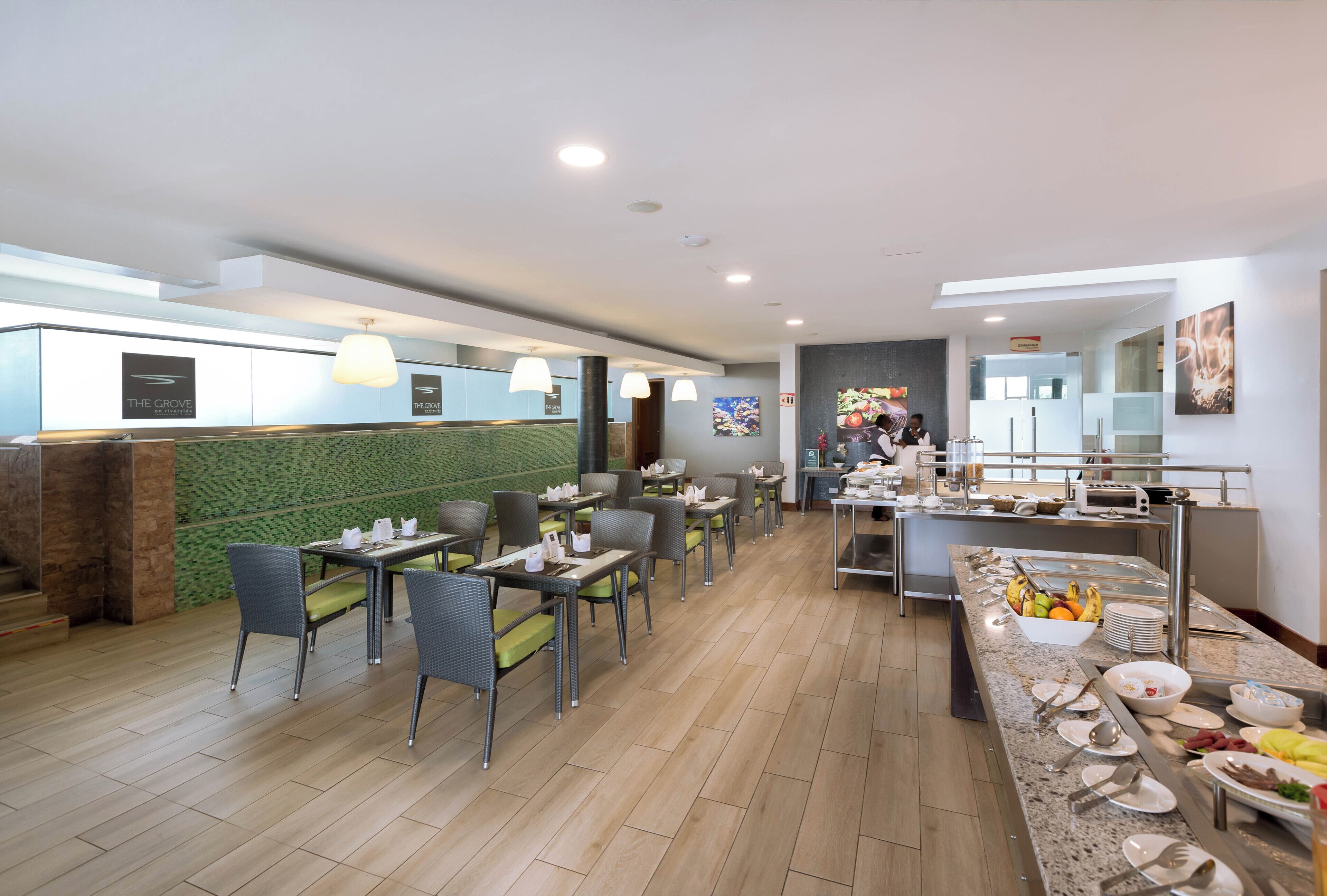 Un restaurante moderno con una pared de mosaicos verdes, mesas de comedor con sillas y una zona de bufé con comida y utensilios.