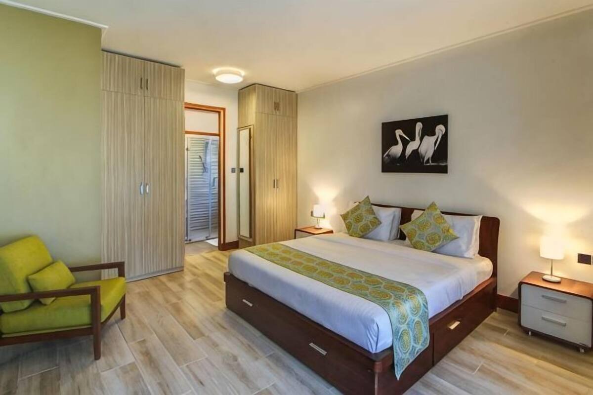 Un dormitorio con cama, mesitas de noche, una silla, un armario y una puerta que da a otra habitación.