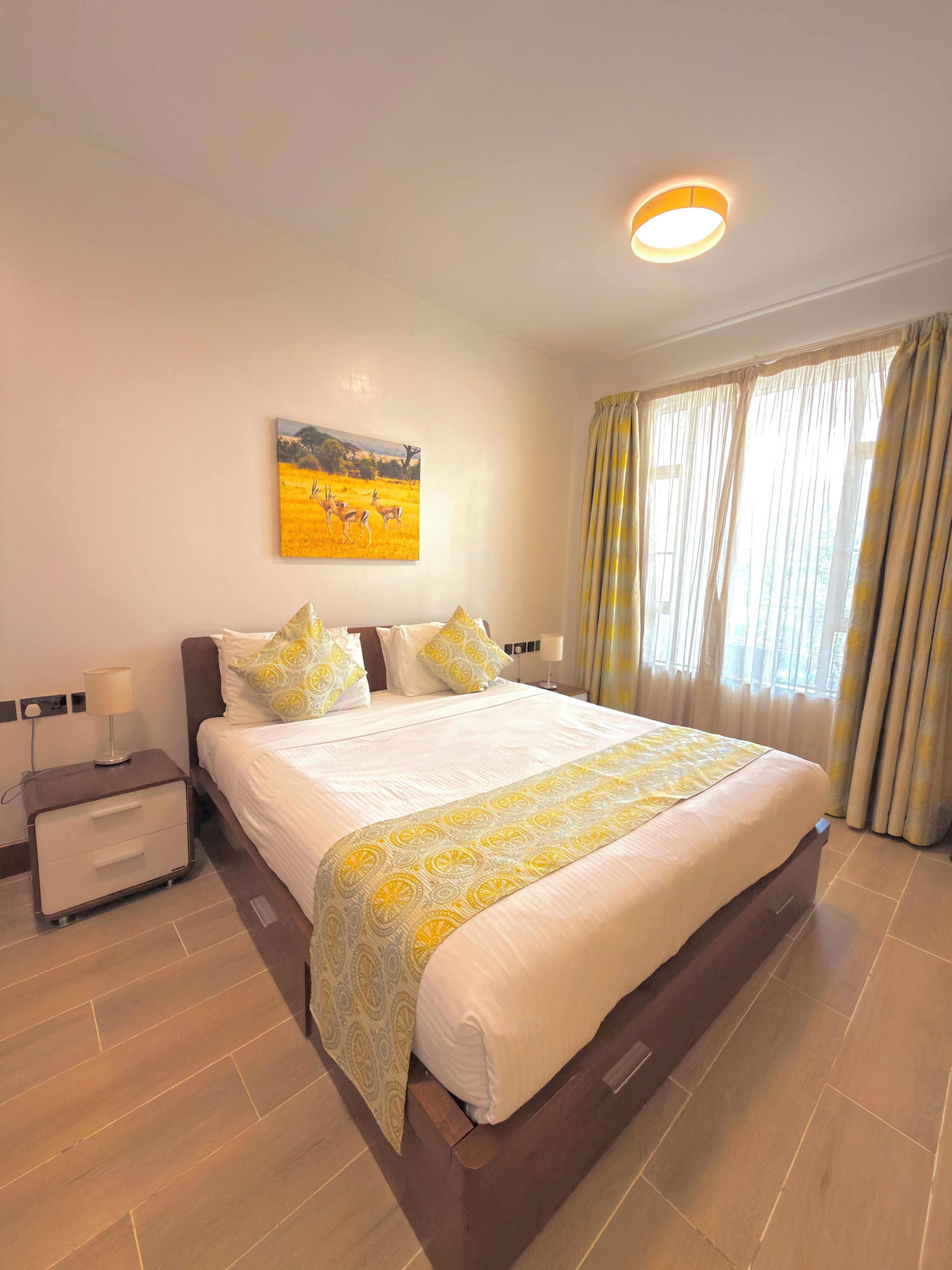 A hotel room with a large bed, a bedside table with a lamp, a painting on the wall, and a window with curtains.
