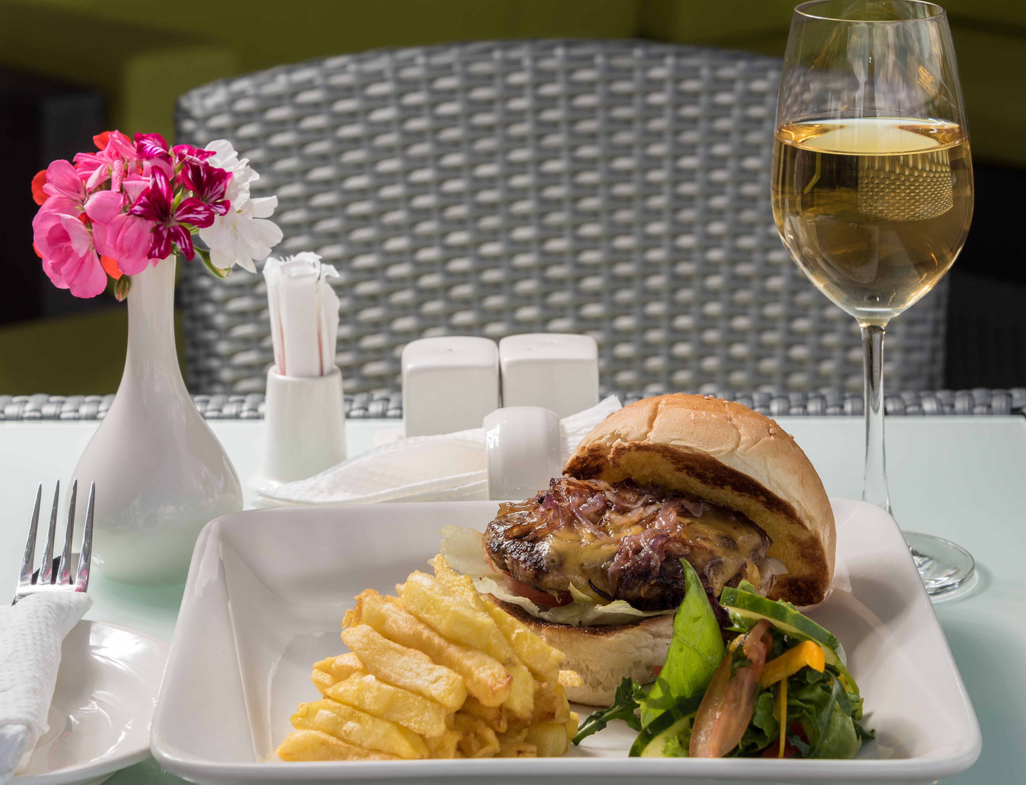 Ett matbord med en smörgås, pommes frites, en sallad, ett glas vitt vin, en vas med blommor, salt- och pepparkar samt en gaffel på en servett.